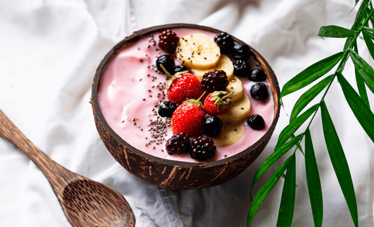 L'image présente un bol de smoothie garni de graines de chia, de bananes tranchées, de fraises coupées et de granola. À côté du bol, on peut voir des fraises et fruits rouge et des bananes sur une surface en bois. 